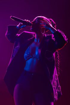 a woman singing into a microphone while standing in front of a purple light with her hands on her head