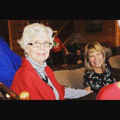 two women sitting on a couch smiling at the camera with other people in the background