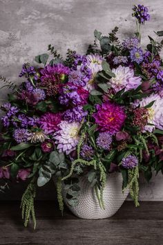 a white vase filled with lots of purple flowers on top of a wooden table next to a wall