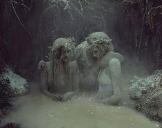 three statues in the middle of a body of water surrounded by trees and bushes, with fog coming from behind them