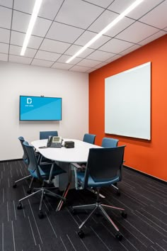 an empty conference room with orange walls and blue chairs around a white table in front of a flat screen tv