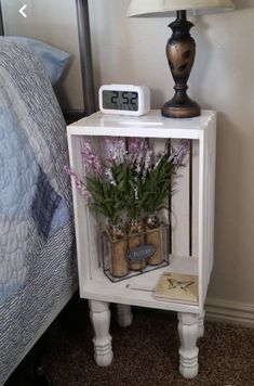 a small white shelf with flowers on it next to a night stand and a lamp