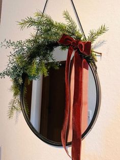 a round mirror hanging on the wall with a red ribbon tied around it and evergreen branches