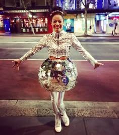 a woman standing on the sidewalk in front of a disco ball costume and holding her arms out