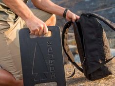 a man holding a black backpack and carrying it on his back while sitting down in the grass