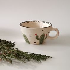 a cactus print coffee cup next to a sprig of pine needles on a white surface