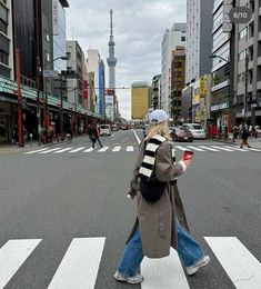 a woman walking across a cross walk holding a cell phone in one hand and a cup in the other