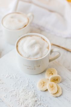 two cups of hot chocolate and sliced bananas on a white counter top with powdered sugar