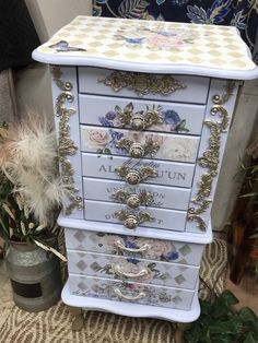 an ornate white and gold chest of drawers with flowers on the top, next to a potted plant