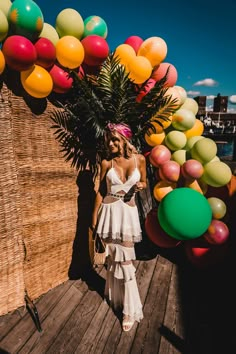 a woman in a white dress standing on a wooden dock with balloons and palm trees