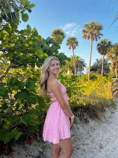 a woman in a pink dress standing on the beach with palm trees and bushes behind her