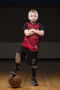 a young boy standing next to a basketball on top of a hard wood floor with his arms crossed