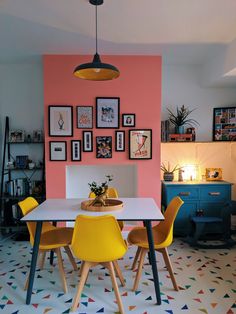 a dining room table with yellow chairs in front of a pink wall and pictures on the wall