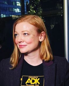 a woman with blonde hair wearing a black shirt and smiling at the camera while sitting in front of a window