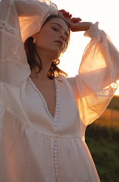 a woman wearing a white dress and holding her hands on her head with the sun shining behind her