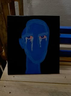 a painting of a blue man with tears on his face and eyes, sitting on a wooden table