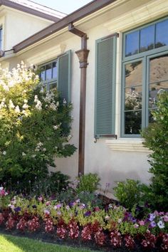 a white house with green shutters and flowers in the front yard, next to a lamp post