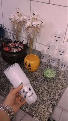 a person holding a coffee cup in front of some cups on a counter top with halloween decorations