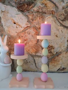 two candles are sitting next to each other on a shelf near a stone wall and bunny figurines