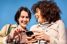 two women are smiling and looking at their cell phones together, one is holding the phone in her hand