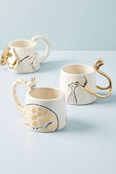 three coffee mugs with designs on them sitting on a blue counter top, one has a gold handle and the other has a giraffe