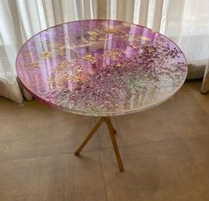 a glass table with purple flowers on it in front of a white curtained window