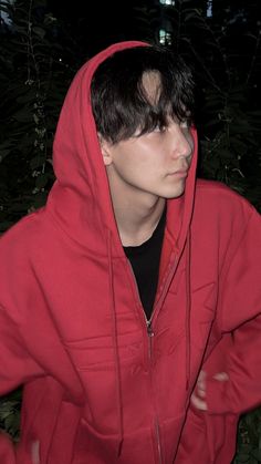 a young man wearing a red hoodie in front of some bushes and trees at night