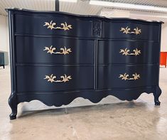 a black dresser with gold leaf handles in a warehouse or office building, it is dark blue and has golden trim on the drawers