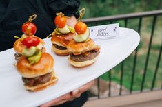 three mini burgers with tomatoes, lettuce and pickles on them sitting on a white platter