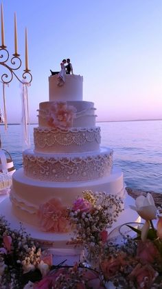 a wedding cake sitting on top of a table next to the ocean with candles in the background