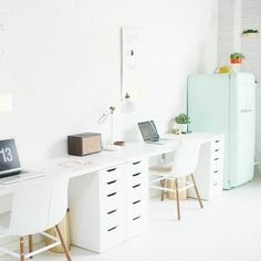 a white desk with two laptops on it and a refrigerator in the back ground