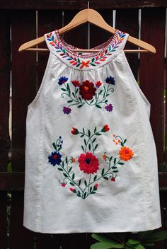 a white top with colorful flowers on it hanging from a wooden hanger next to a fence