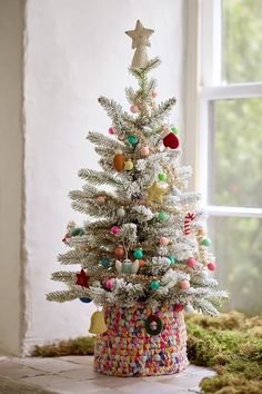 a small white christmas tree with colorful ornaments and decorations on it's base in front of a window