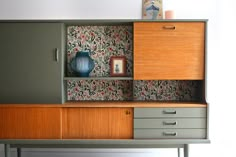 an old dresser with drawers and a vase on it's top, in front of a wallpapered background