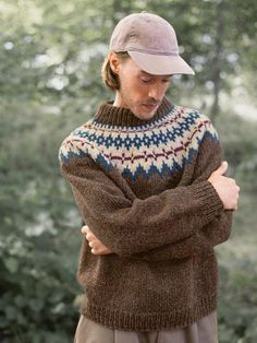 a woman standing in front of trees wearing a brown sweater and hat with her arms crossed