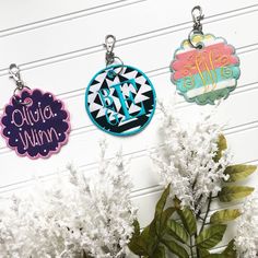 three personalized keychains are shown next to some white flowers and plants in front of a wall