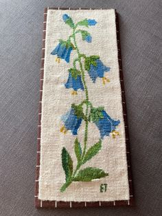 a cross - stitch bookmark with blue flowers and green leaves on white fabric, sitting on a gray surface