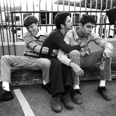 three young men sitting on a bench in a parking lot, one is holding his arm around the other's neck