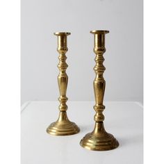 two brass candlesticks sitting side by side on a white surface with one candle in the middle