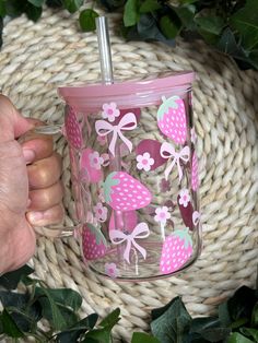 a hand holding a pink cup with strawberries and bows on it next to some green leaves