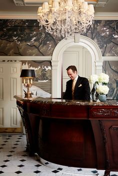 a man in a suit standing at the front desk of a hotel lobby with chandelier