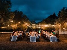 a group of people sitting at tables in the middle of a field with lights strung over them