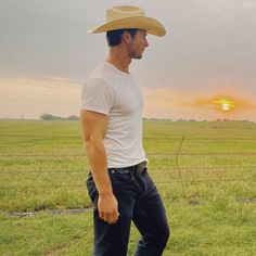 a man wearing a cowboy hat standing in front of a fence with the sun setting behind him