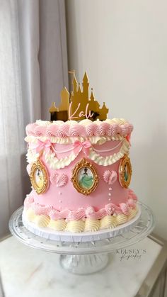 a pink cake with gold decorations on top sitting on a white table in front of a window