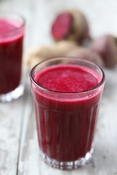 two glasses filled with red liquid sitting on top of a wooden table next to fresh vegetables