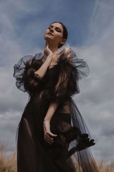 a woman standing in a field wearing a black dress and holding her hand to her face