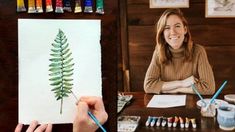 a woman sitting at a table holding a pencil and drawing a fern leaf on paper