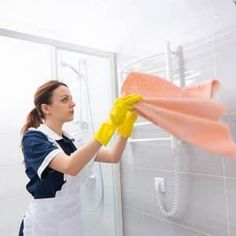 a woman in an apron cleaning a shower wall with a rag and yellow rubber gloves