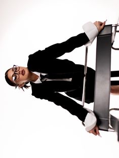 a woman in black suit and glasses leaning against a chair