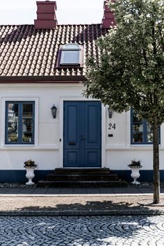 a white house with blue shutters and a tree in front of it on a cobblestone street